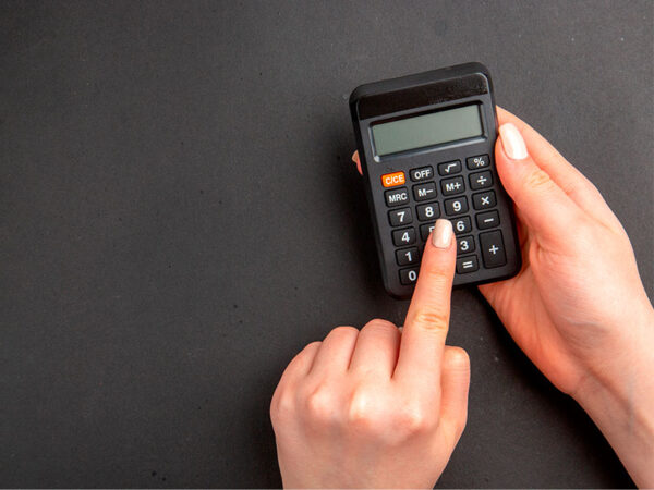 A imagem, de fundo grafite, mostra a mão de uma mulher de pele clara segurando uma calculadora pequena preta. Com a outra mão, ela digita um número no aparelho.