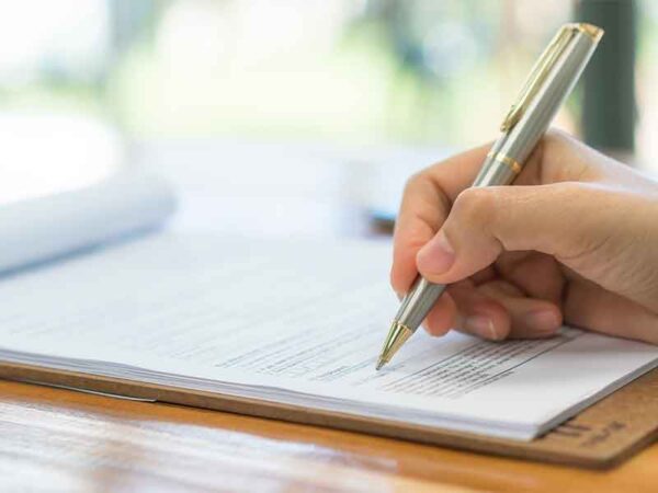 A imagem mostra uma mão segurando uma caneta dourada prestes a assinar um documento em uma pilha de papeis sobre uma mesa de madeira envernizada. No fundo é possível notar um ambiente arborizado, iluminado pelo sol.