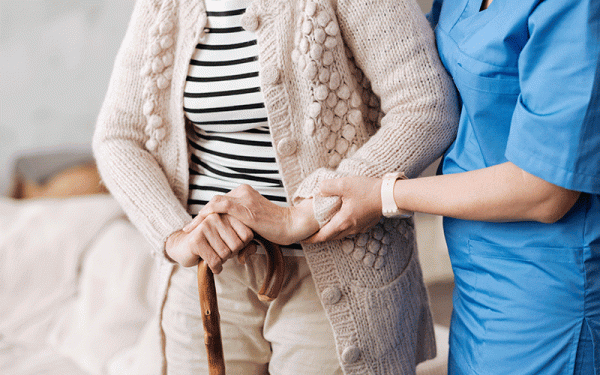 Uma idosa vestindo casaco segura uma bengala para se locomover. Ela conta o auxílio das mãos de uma enfermeira, que está vestida com uniforme da cor azul.