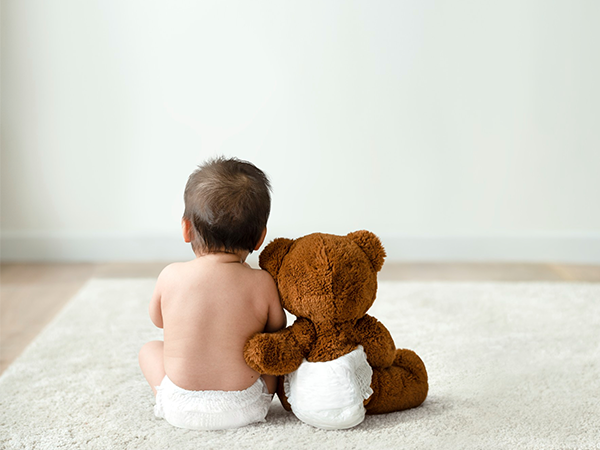 A foto mostra um bebê e um urso marrom, sentados de costas para a imagem, sob um tapete de pelos beges. Ambos vestem uma fralda branca e estão sentados lado a lado. O braço esquerdo do urso está apoiado nas costas do bebê. Ao fundo é possível visualizar uma parede branca.