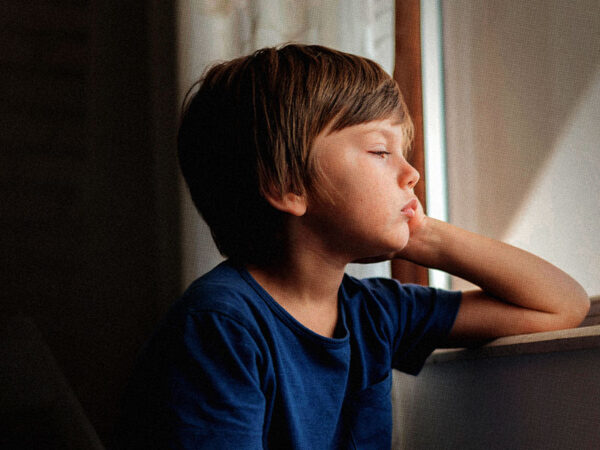 Imagem de menino de pele bronzeada, cabelos curtos, lisos e loiros escuros. Ele está sentado de frente à uma janela, segura o rosto com uma das mãos e tem um semblante desanimado.